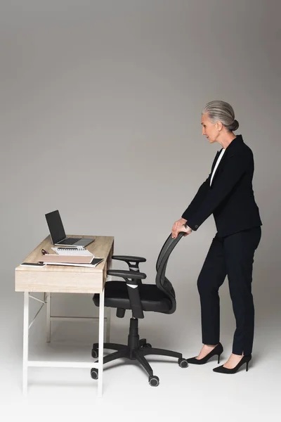 Vista lateral de la mujer de negocios madura de pie cerca de la silla de oficina y mesa de trabajo sobre fondo gris - foto de stock