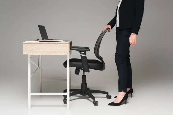 Обрезанный вид деловой женщины, стоящей возле офисного стула и ноутбука на столе на сером фоне — стоковое фото