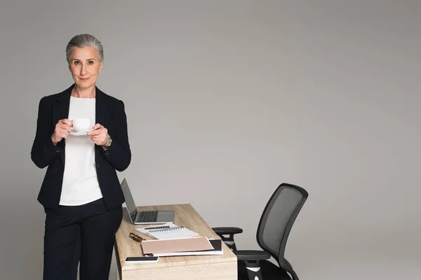 Madura mujer de negocios sosteniendo taza cerca de papeles y gadgets en la mesa aislado en gris - foto de stock