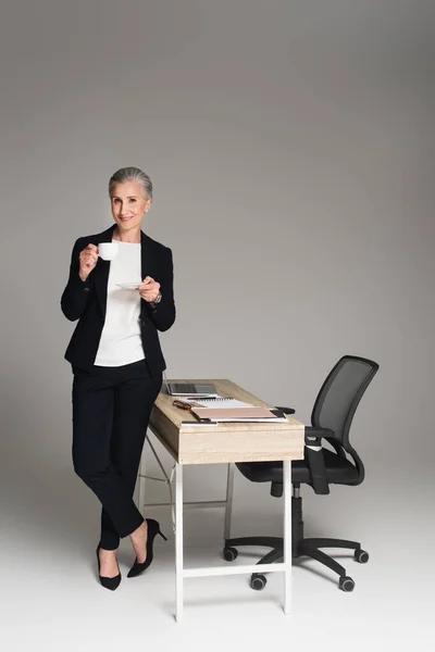 Mujer de negocios sonriente sosteniendo taza cerca de dispositivos y papeles en la mesa sobre fondo gris - foto de stock