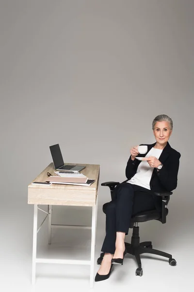 Reife Geschäftsfrau hält Tasse in der Nähe von Gadgets und Papierkram auf dem Tisch auf grauem Hintergrund — Stockfoto