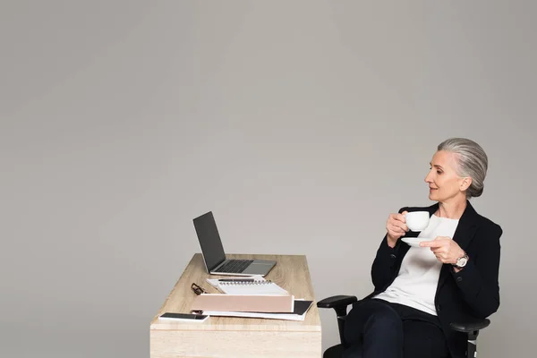 Зріла бізнес-леді з чашкою дивиться на ноутбук біля паперів на столі ізольовано на сірому — стокове фото