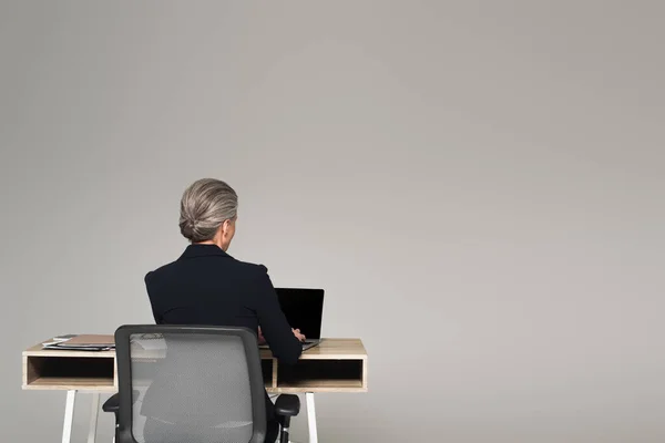 Vista posterior de la mujer de negocios madura usando el ordenador portátil cerca de documentos en la mesa aislado en gris - foto de stock