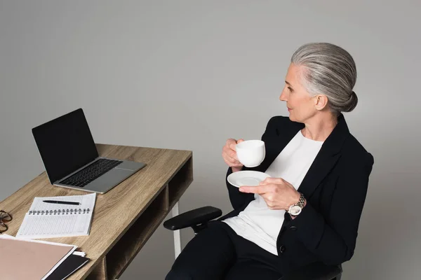 Mujer de mediana edad con taza de café sentado cerca de la computadora portátil y portátil en la mesa aislado en gris - foto de stock