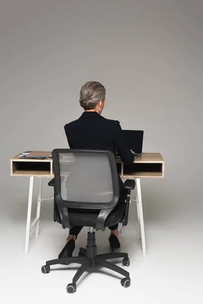 Обратный вид менеджера средних лет с помощью ноутбука рядом с бумажной работой на столе на сером фоне — стоковое фото