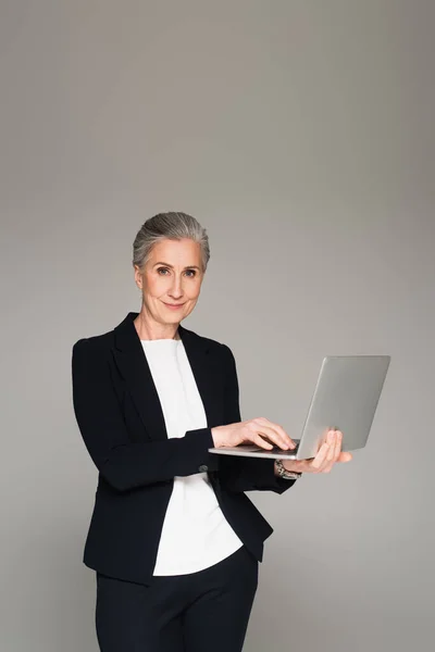 Madura mujer de negocios utilizando el ordenador portátil y mirando a la cámara aislada en gris - foto de stock