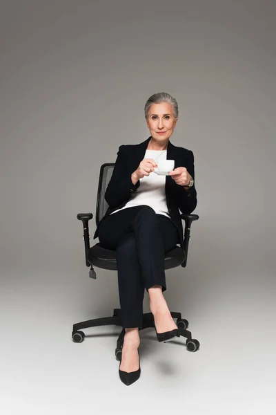 Empresária em uso formal segurando xícara de café na cadeira de escritório em fundo cinza — Fotografia de Stock