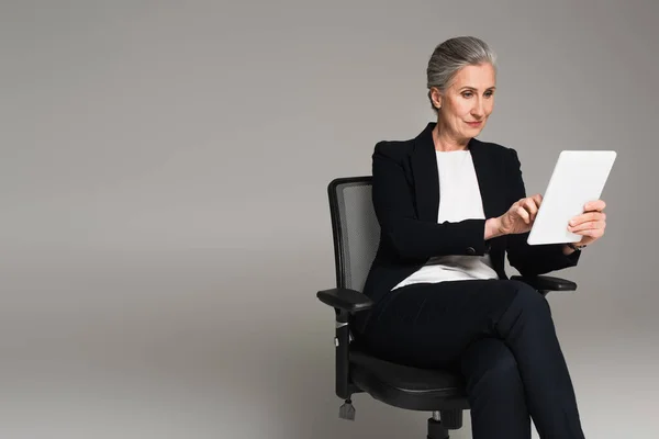 Femme d'affaires aux cheveux gris utilisant une tablette numérique sur chaise de bureau isolée sur gris — Photo de stock