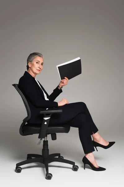 Mature businesswoman holding paper folder on office chair on grey background — Stock Photo