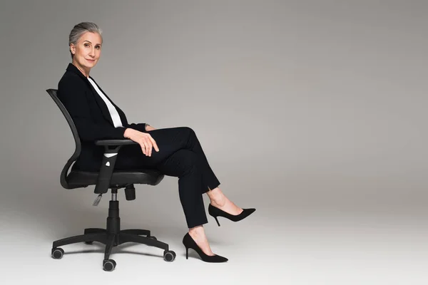 Femme d'affaires d'âge moyen avec jambes croisées regardant caméra sur chaise de bureau sur fond gris — Photo de stock