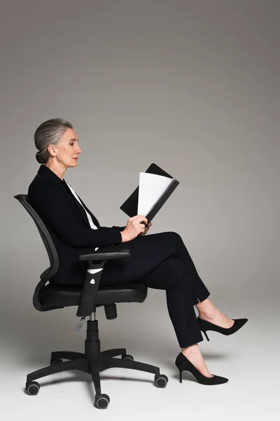 Side view of mature businesswoman looking at documents on office chair on grey background — Stock Photo