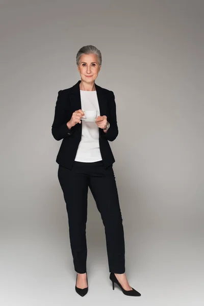 Femme d'affaires senior avec tasse et soucoupe regardant la caméra sur fond gris — Photo de stock