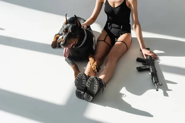Cropped view of woman in bodysuit sitting near rifle and doberman on white floor with grey shadows — Stock Photo