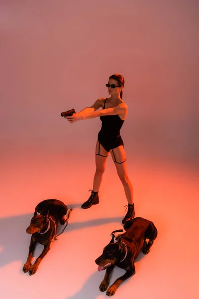 Стильна жінка в боді-костюмі з ручною зброєю поблизу домерзан на рожевому фоні з жовтим світлом — стокове фото