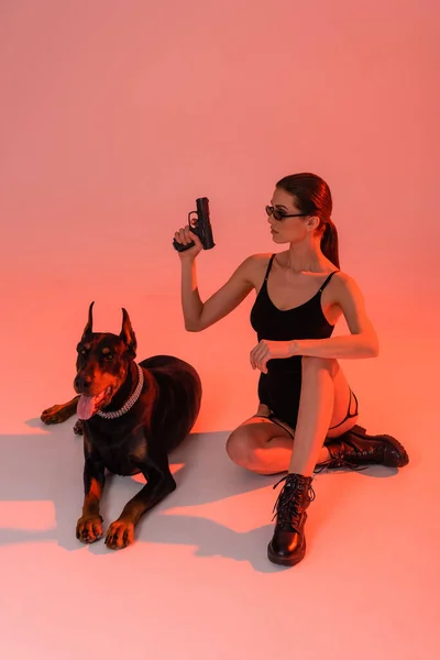 Sexy Frau in Body und Stiefeln mit Pistole in der Nähe von Dobermann auf rosa Hintergrund mit gelbem Licht — Stockfoto