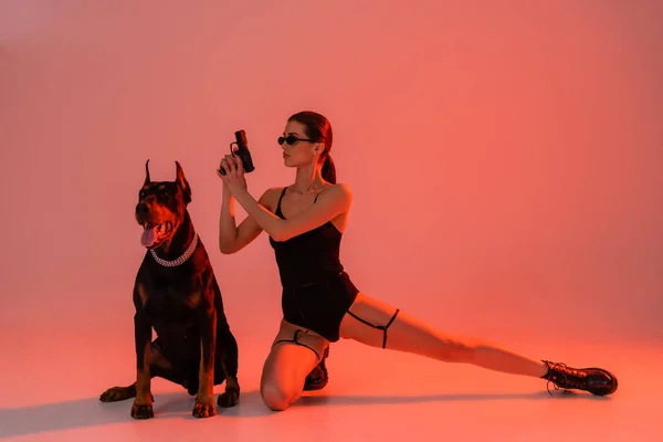 Brunette woman in bodysuit and sunglasses holding gun near doberman on pink background with yellow light — Stock Photo