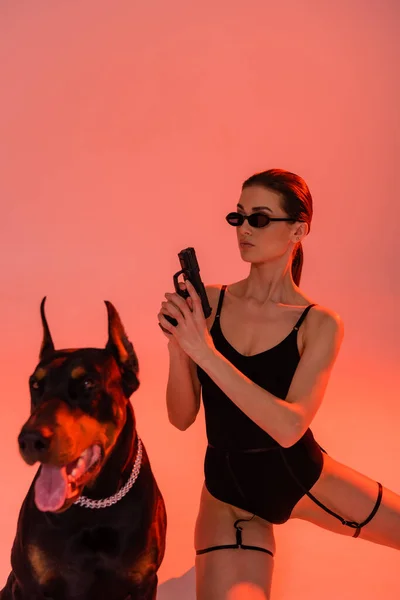 Размытый доберман собака рядом с сексуальной женщиной с пистолетом на розовом фоне с желтым светом — стоковое фото