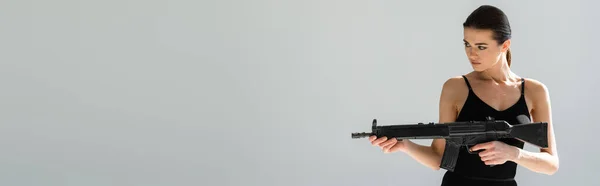 Brunette woman in bodysuit aiming with rifle isolated on grey, banner — Stock Photo