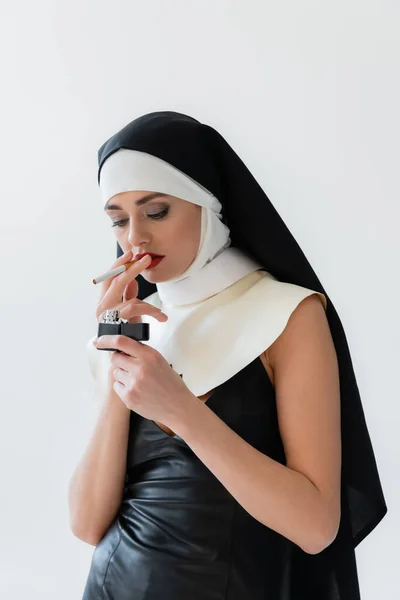 Monja joven en vestido de cuero iluminación cigarrillo aislado en gris - foto de stock