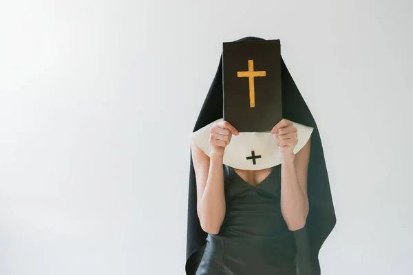 Monja en vestido sexy oscureciendo la cara con la Biblia santa aislado en gris - foto de stock