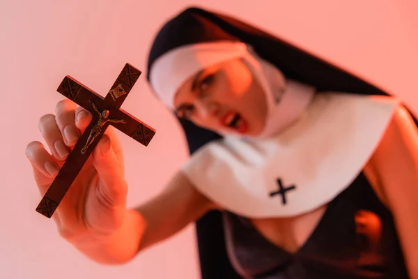 Angry nun holding crucifix while screaming on blurred foreground isolated on pink — Stock Photo