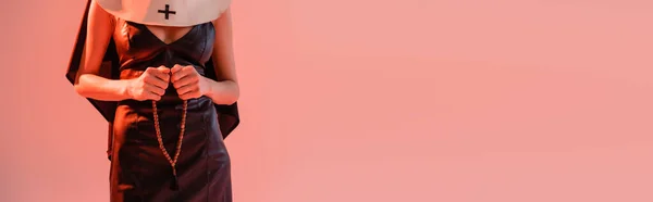 Cropped view of sexy nun in leather dress holding prayer beads isolated on pink, banner — Stock Photo