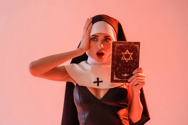 Monja impactada en vestido sexy tocando la cabeza mientras sostiene la biblia judía aislada en rosa - foto de stock
