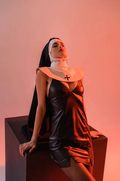 Sensual nun in sexy dress posing with closed eyes near black cube on pink background — Stock Photo