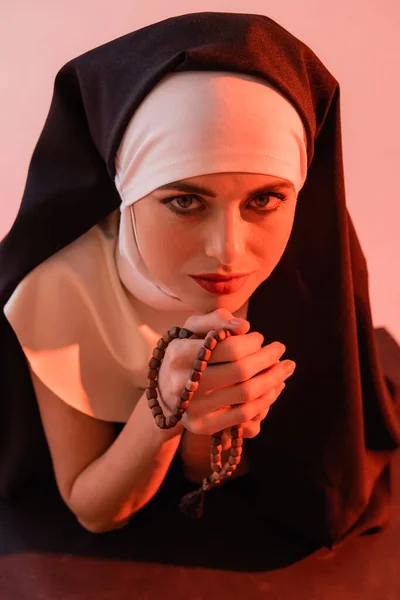 Monja joven mirando a la cámara mientras sostiene rosario aislado en rosa - foto de stock