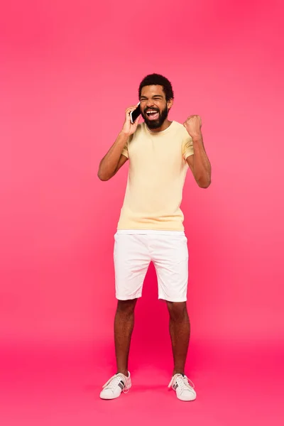 Longitud completa de hombre afroamericano emocionado hablando en el teléfono inteligente en rosa - foto de stock