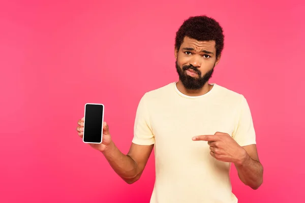 Спантеличений афроамериканський чоловік вказує на смартфон з порожнім екраном ізольовано на рожевому — стокове фото