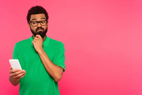 Hombre afroamericano pensativo en camiseta verde usando teléfono inteligente aislado en rosa - foto de stock