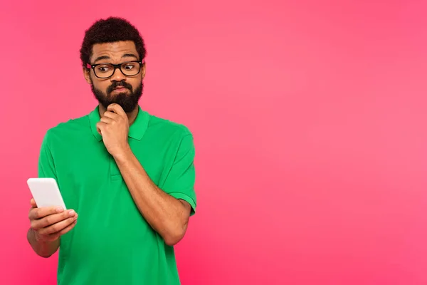 Pensive african american man in green t-shirt using smartphone isolated on pink — Stock Photo