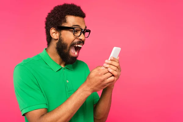 Excited african american man in green t-shirt using smartphone isolated on pink — Stock Photo