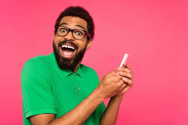 Asombrado hombre afroamericano en camiseta verde usando teléfono inteligente aislado en rosa - foto de stock