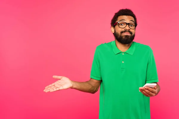 Verwirrter afrikanisch-amerikanischer Mann in grünem T-Shirt hält Smartphone isoliert auf rosa — Stockfoto