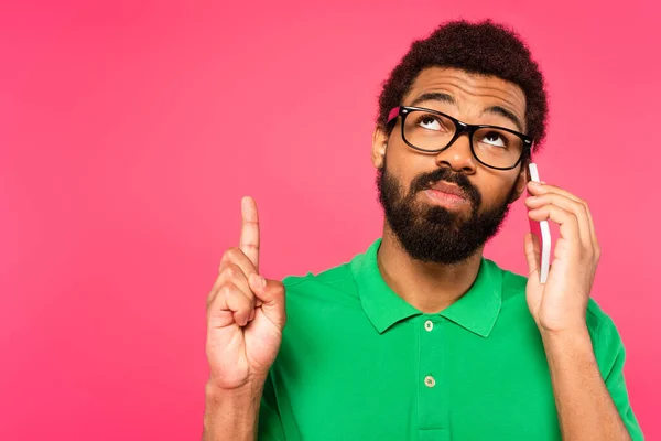 Africano americano hombre en gafas hablando en smartphone y apuntando hacia arriba aislado en rosa - foto de stock