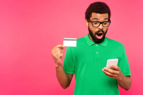 Sorprendido hombre afroamericano en gafas usando teléfono inteligente y la celebración de la tarjeta de crédito aislado en rosa - foto de stock