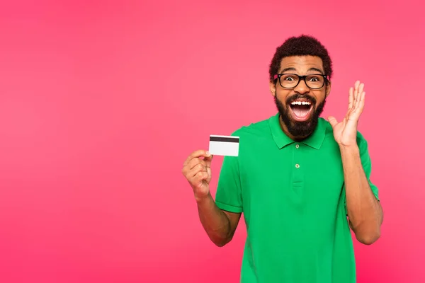 Étonné homme afro-américain dans des lunettes tenant la carte de crédit isolé sur rose — Photo de stock