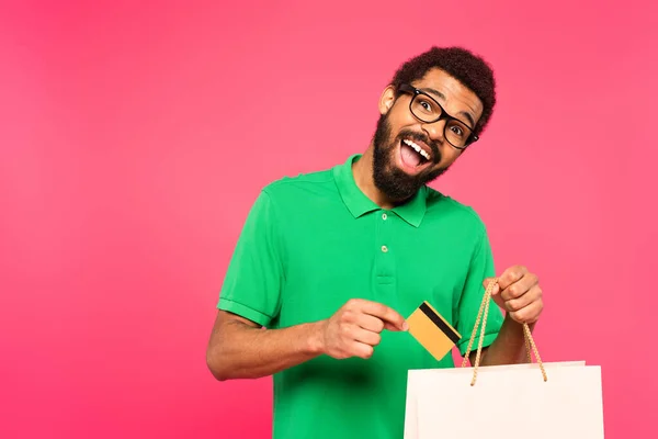 Homem americano africano espantado em óculos segurando cartão de crédito perto de saco de compras isolado em rosa — Fotografia de Stock