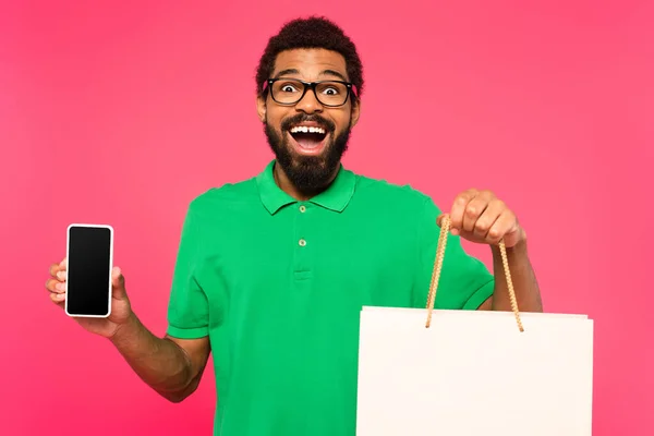 Здивований афроамериканський чоловік в окулярах тримає сумку для покупок і смартфон з порожнім екраном ізольовано на рожевому — стокове фото
