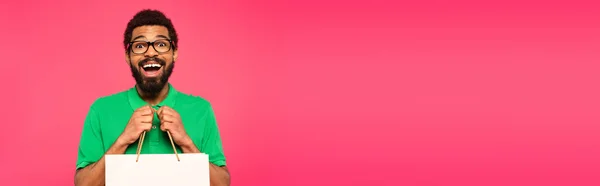 Uomo afroamericano stupito in occhiali con borsa della spesa isolata su rosa, banner — Foto stock