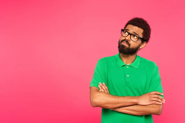 Hombre afroamericano pensativo en gafas mirando hacia otro lado mientras está de pie con los brazos cruzados aislados en rosa - foto de stock