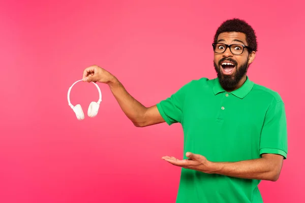 Homem americano africano feliz em óculos apontando para fones de ouvido sem fio isolados em rosa — Fotografia de Stock