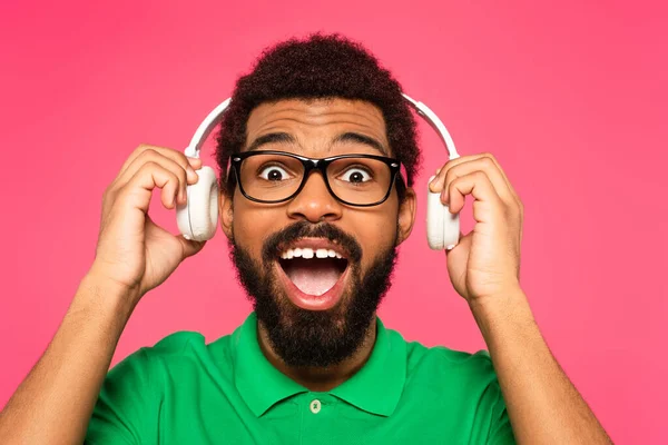 Aufgeregter afrikanisch-amerikanischer Mann in Brille mit drahtlosen Kopfhörern isoliert auf rosa — Stockfoto