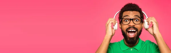 Возбужденный африканский американец в очках держа беспроводные наушники изолированы на розовый, баннер — стоковое фото