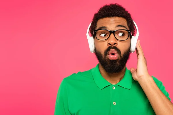 Chocado afro-americano homem em óculos e fones de ouvido sem fio isolado em rosa — Fotografia de Stock