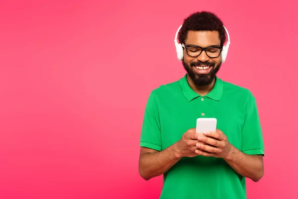 Happy african american man in glasses and wireless headphones using smartphone  on pink — Stock Photo