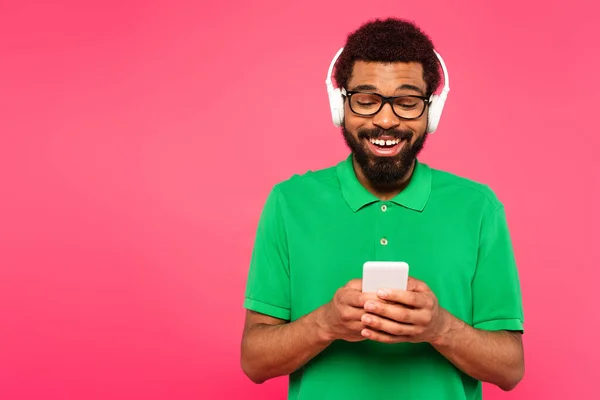 Fröhlicher afrikanisch-amerikanischer Mann mit Brille und drahtlosen Kopfhörern, Smartphone isoliert auf rosa — Stockfoto