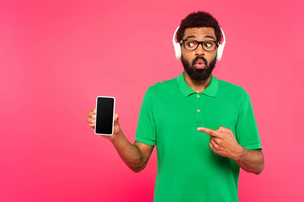 Chocado homem americano africano em fones de ouvido sem fio apontando para smartphone isolado em rosa — Fotografia de Stock